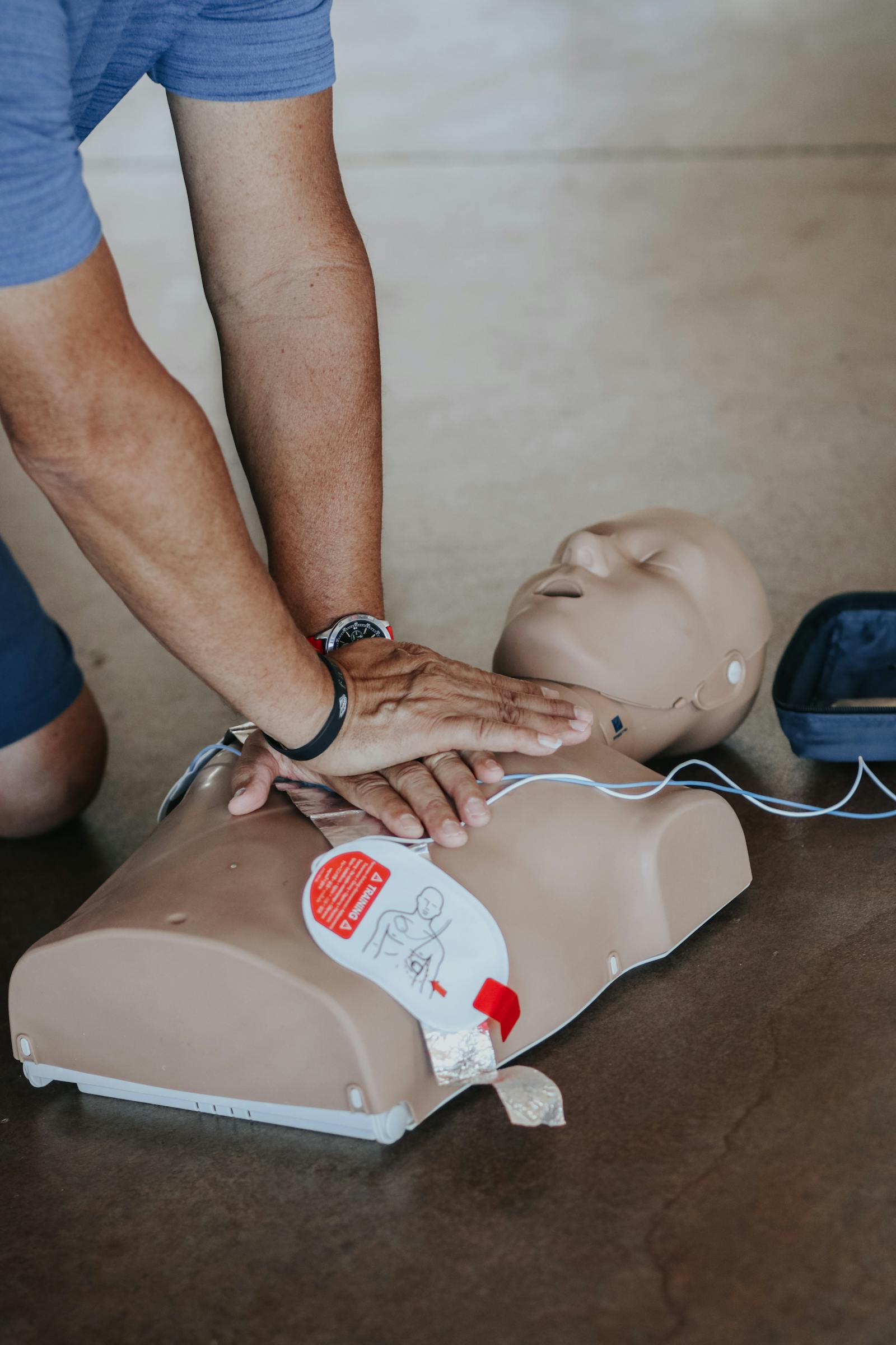 Hands performing chest compressions on a CPR mannequin with AED pads attached.